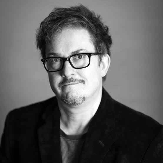A black and whit image of a man with tousled hair and moustace and light beard he is smiling with his mouth closed. He wears dark framed glasses