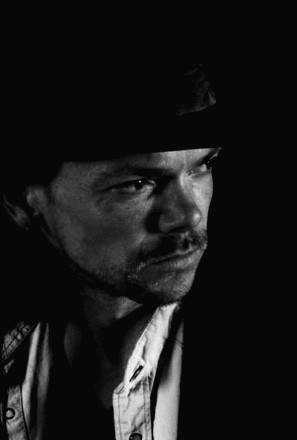 a black and white image of man with dark hair and moustache his eyes are shining and we can see him almost in profile. He is wearing a hat of some kind.