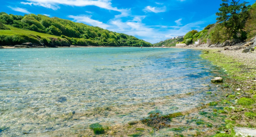 a shallow river stretching into a narrow valley with green sides. There are pebbles under the water which is turquoise under a blue sky.